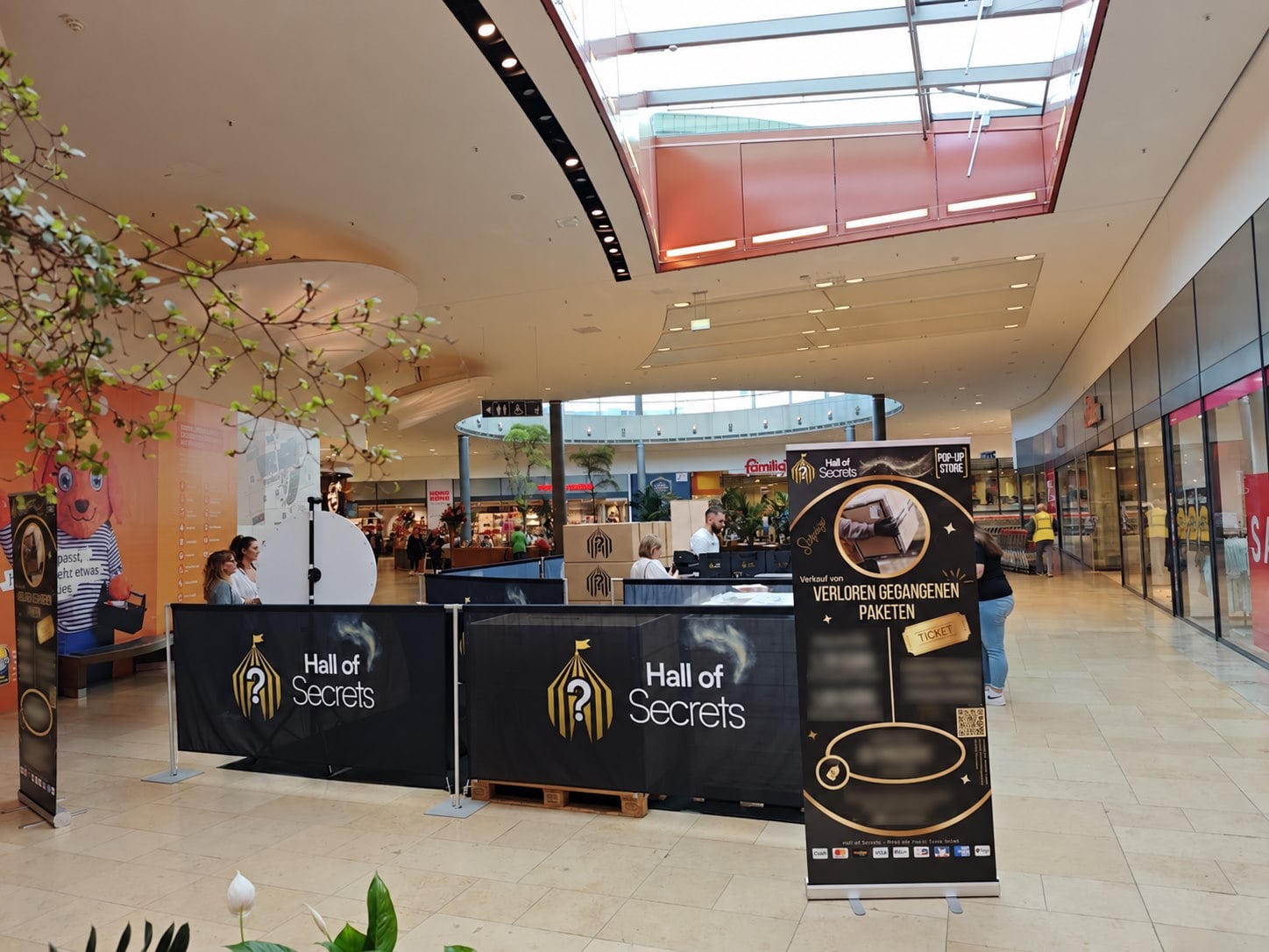 Der Hall-of-Secrets-Stand im Einkaufscenter mit schwarz-goldenen Bannern und großen Hall-of-Secrets-Logos.